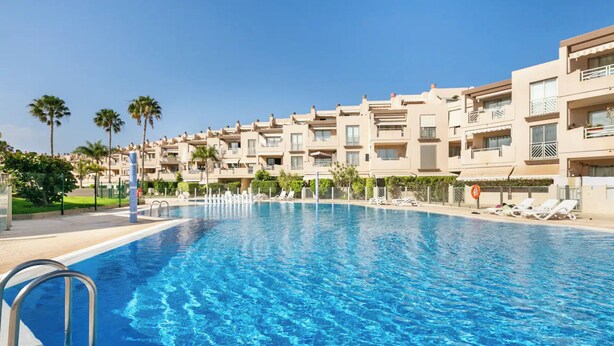 A swimming pool with clear blue water, surrounded by multi-story buildings with balconies and a clear sky.