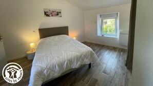 A bedroom with a bed, a nightstand, a window, and a wall-mounted picture. - Cozy 6-Person Cottage in Chanverrie with Private Bathrooms, Kids' Play Area, and Terrace (Chanverrie)
