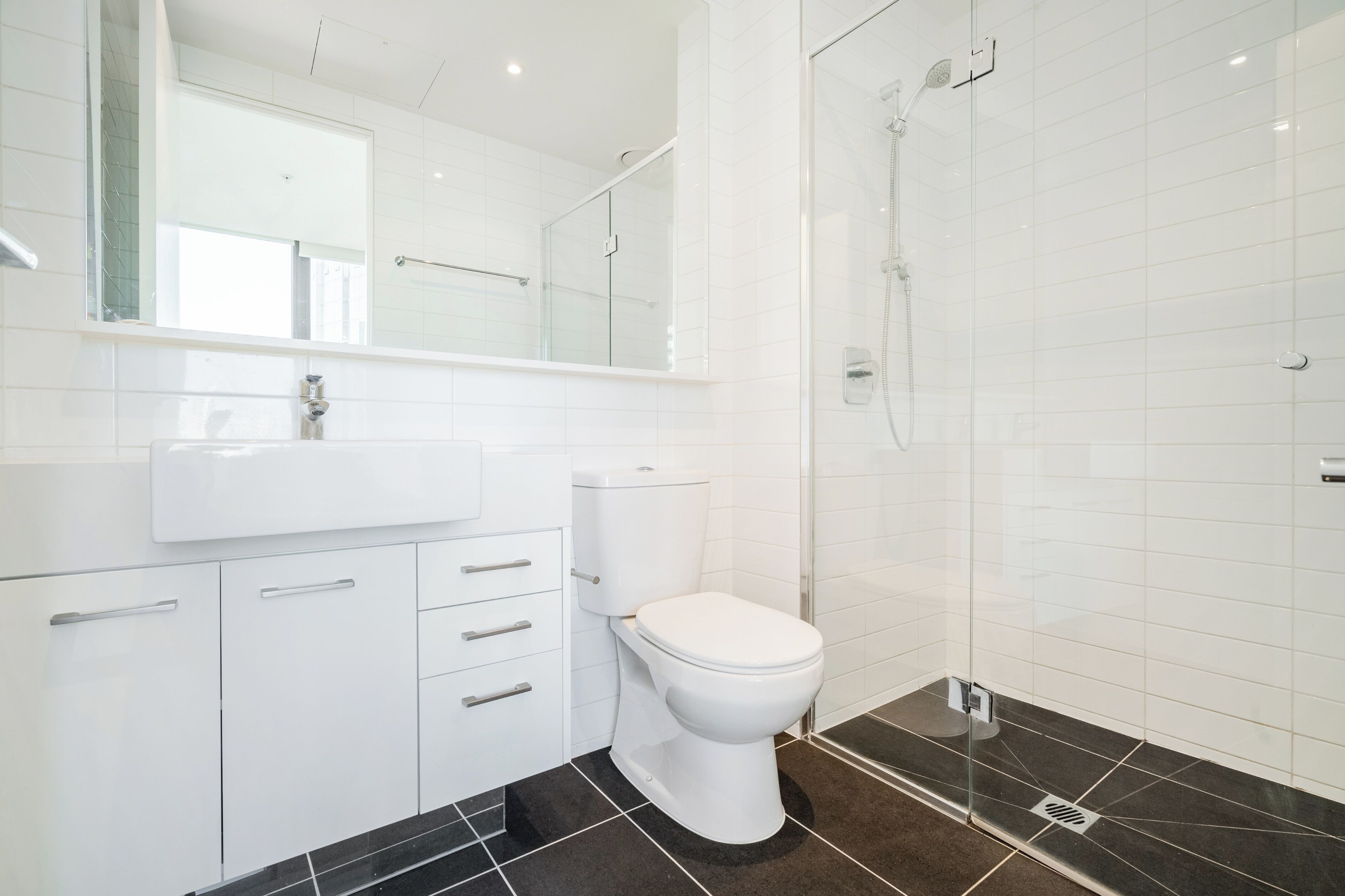 Un baño moderno con inodoro blanco, cabina de ducha con paredes de vidrio y suelo de baldosas negras.