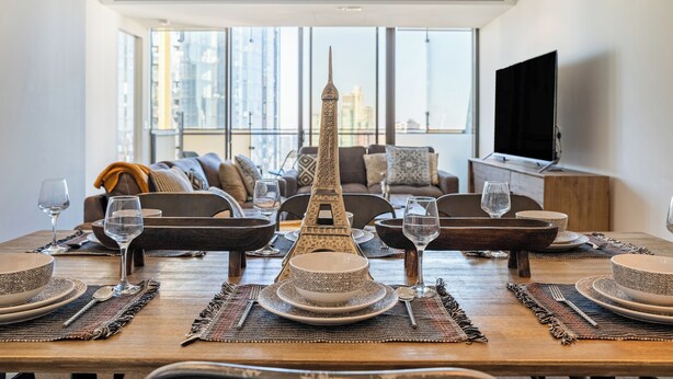 Un coin repas avec une table dressée pour quatre personnes, une tour Eiffel décorative en guise de centre de table, et un salon moderne en arrière-plan, avec une grande fenêtre et un téléviseur à écran plat.