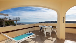 A balcony with a pool chair, a table with chairs, and a view of the city and sea.