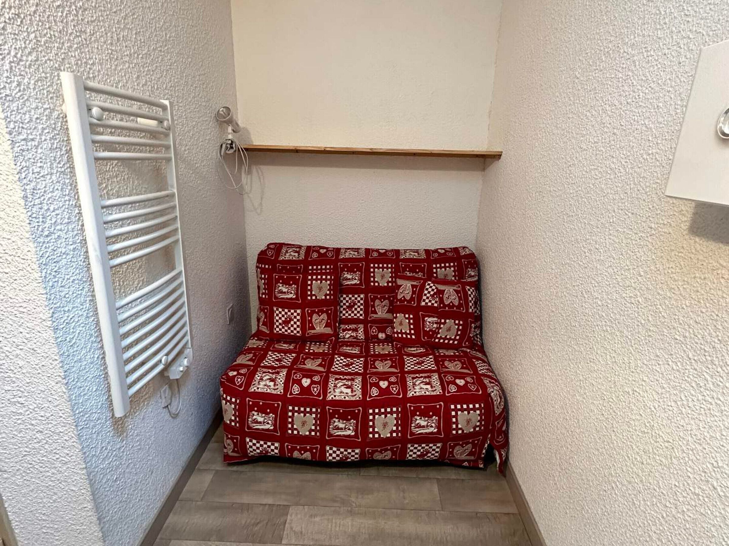 A small, narrow room with a radiator on the left, a wooden shelf above the bed, and a white wall.