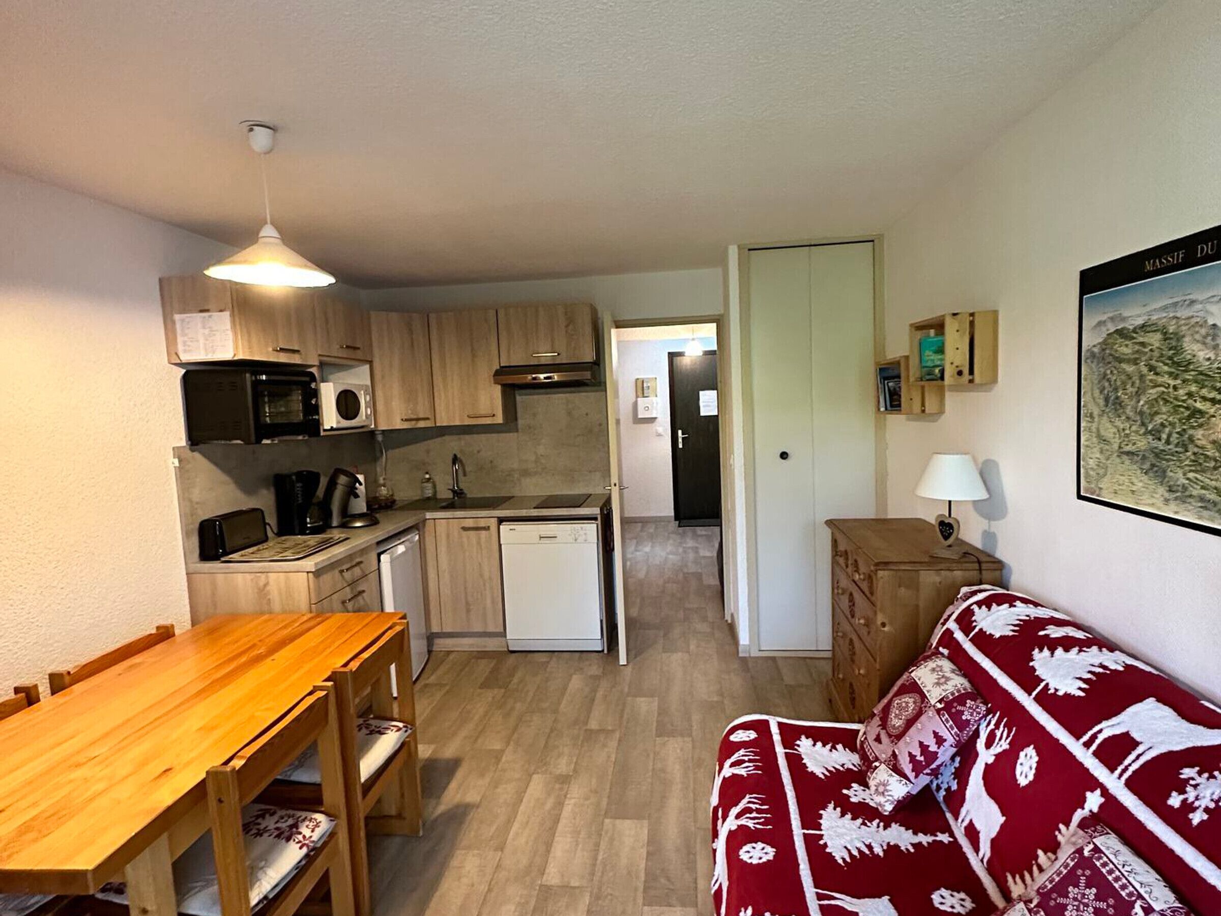 A compact kitchen with wooden cabinets, a microwave, and a dishwasher. There is a wooden dining table with chairs. A red patterned sofa is against the wall.