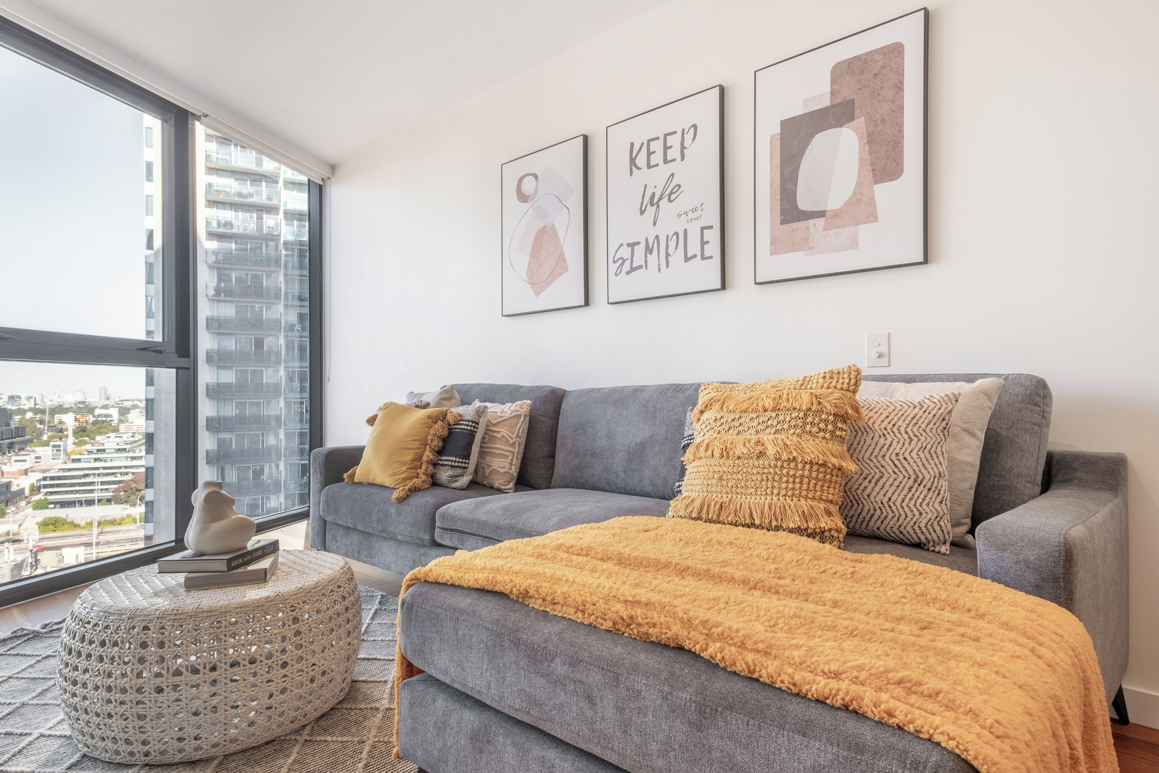 A modern living room with a grey sofa, a yellow throw blanket, and a round ottoman. There are three framed artworks on the wall and a view of a cityscape through a large window.