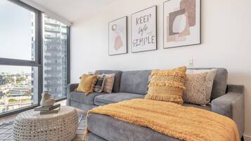 A modern living room with a grey sofa, a yellow throw blanket, and a round ottoman. There are three framed artworks on the wall and a view of a cityscape through a large window.