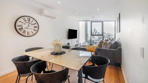 A modern living room with a dining area, a large city view, and a wall-mounted clock. - Mega Style Apartments Bella (Southbank)