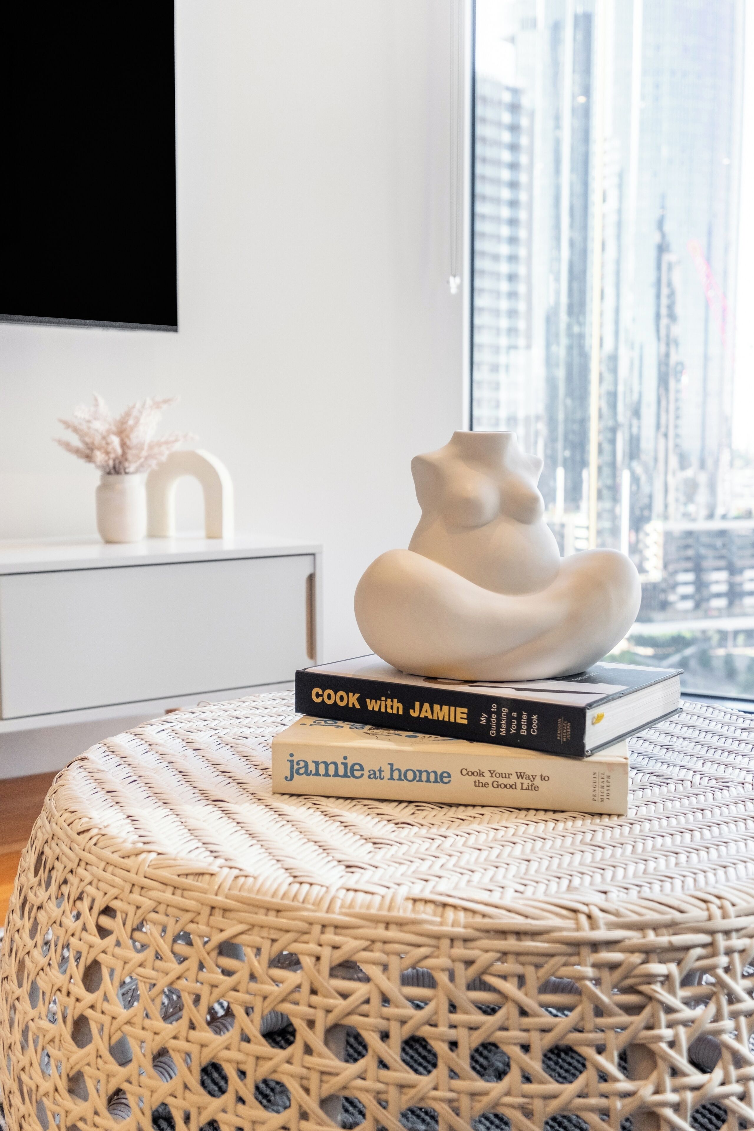 A modern living room with a white sofa, a decorative vase, and two cookbooks on a woven ottoman.