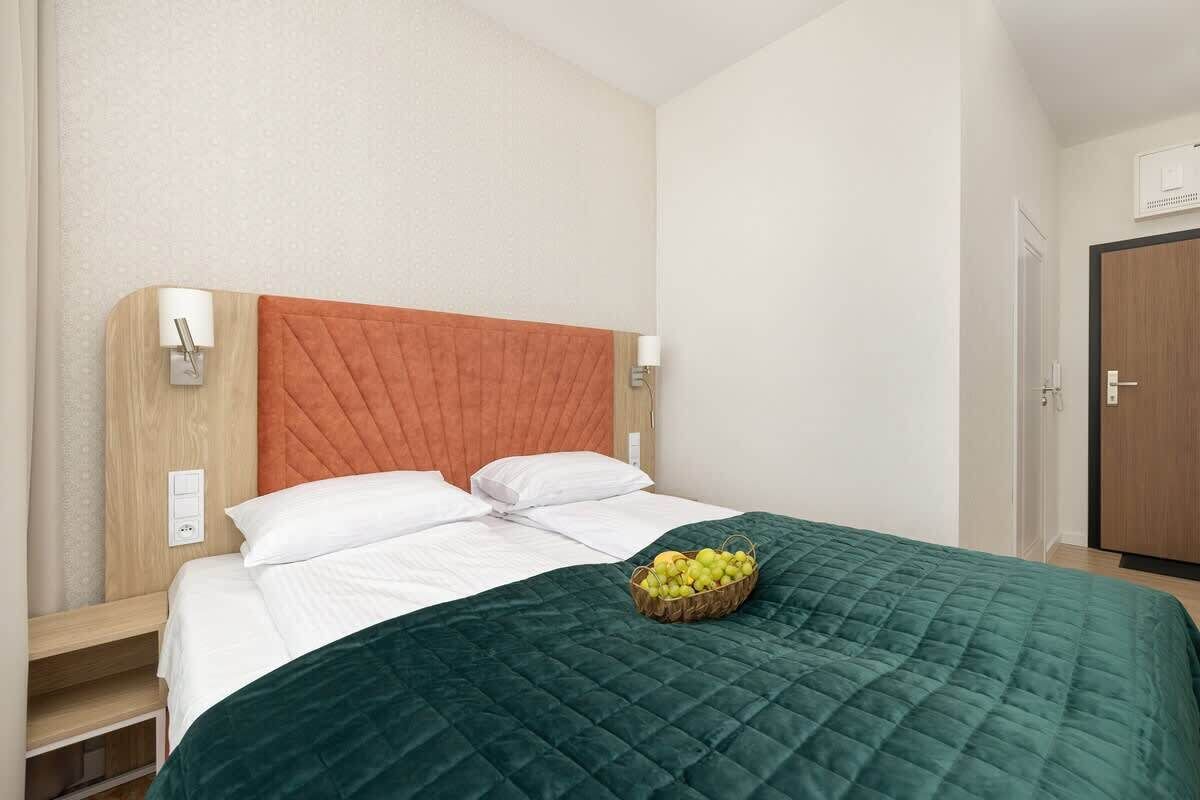 Quarto de hotel com uma cama, cabeceira de madeira, colcha verde acolchoada e uma cesta de frutas na cama.