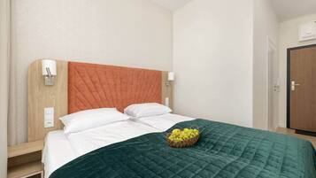 Quarto de hotel com uma cama, cabeceira de madeira, colcha verde acolchoada e uma cesta de frutas na cama.