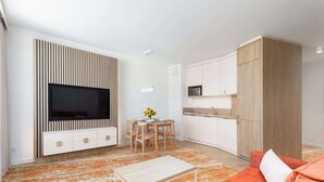 A modern living room with a flat-screen TV, a dining table with chairs, a kitchen area with cabinets, and a sofa. - Modern Apartment with Balcony | Parking, 450 m to the Beach (Miedzyzdroje)