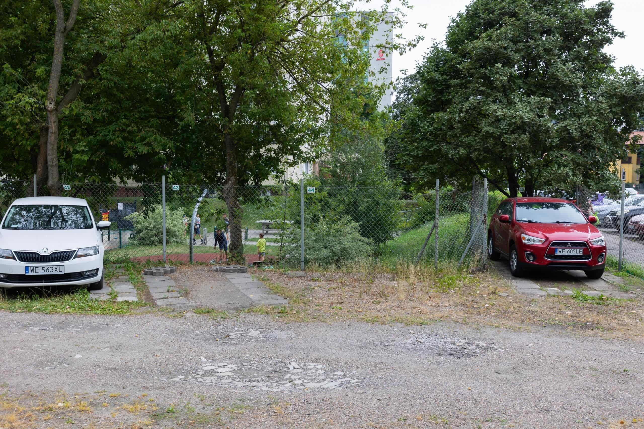 A parking area with two cars, a white car and a red car, parked on a gravel surface. There are trees and a fence in the background.