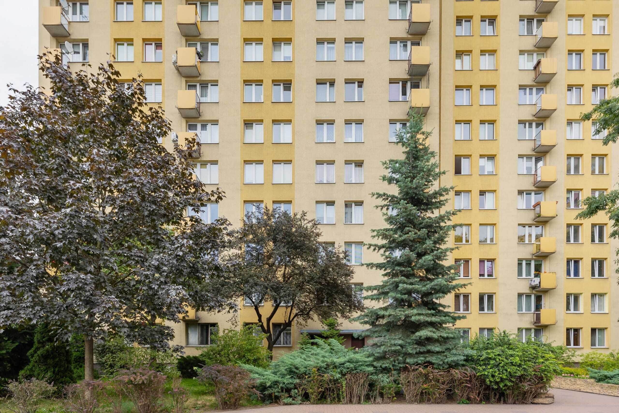 A multi-story residential building with balconies, surrounded by trees and shrubs.