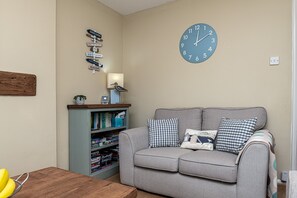 Una sala de estar con un sofá gris, almohadones de cuadros, una estantería, un reloj de pared y una mesa de madera con plátanos.