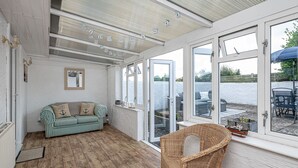 A sunlit room with a wooden floor, a striped sofa, a wicker chair, and large windows offering a view of the outdoors. - Tan Y Wylan -  a bungalow that sleeps 6 guests  in 3 bedrooms (Rhosneigr)