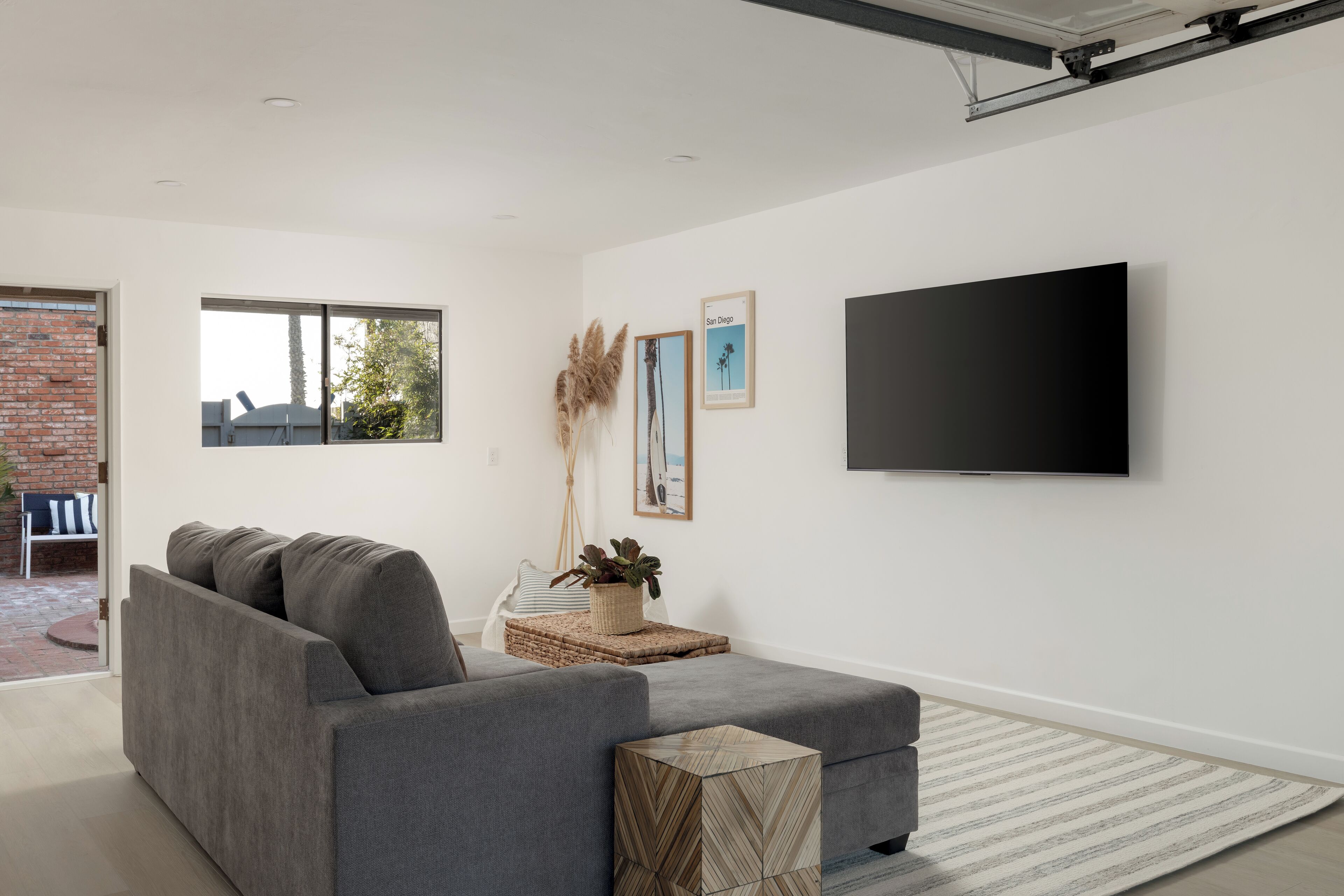 A modern living room with a grey sofa, a flat-screen TV mounted on the wall, and a large window providing natural light.