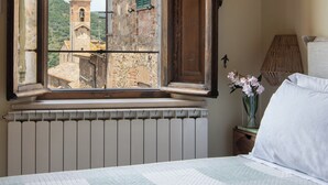 Ein Schlafzimmer mit Bett, einem Fenster mit geöffnetem Klappladen, einem Heizkörper, einer Lampe und einer Vase mit Blumen.