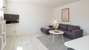 Uma sala de estar moderna com um sofá cinzento, uma mesa de centro branca e uma televisão de tela plana na parede.