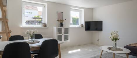 Una sala de estar moderna con comedor, un televisor montado en la pared y un ventanal con vistas a un edificio.