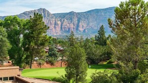 Un campo de golf con montañas al fondo, rodeado de árboles y un edificio con techo inclinado.