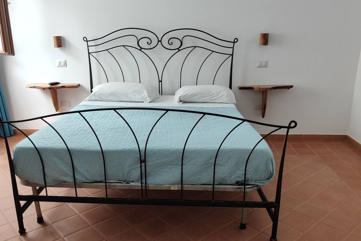 Una cama con estructura metálica, dos mesitas de noche de madera y una colcha azul claro.