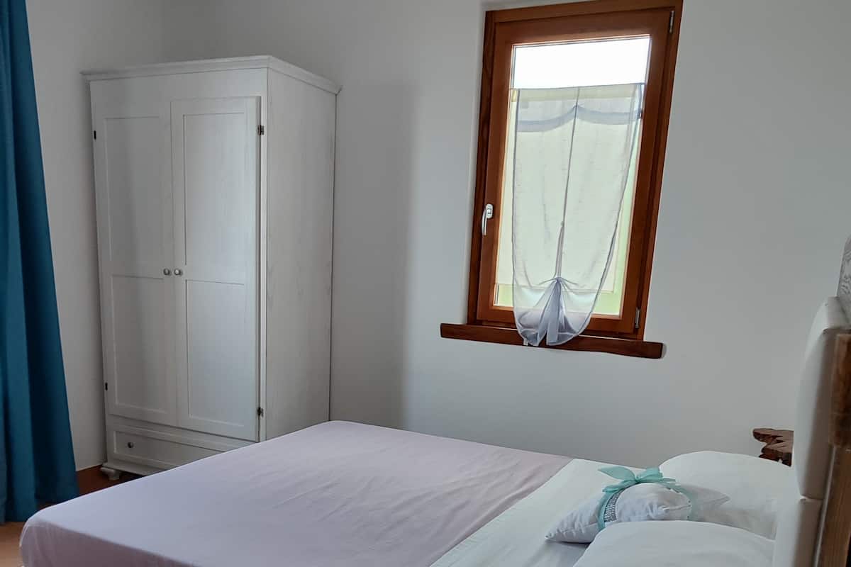 Un dormitorio con cama, armario, ventana con cortinas y una mesa de madera con un control remoto.