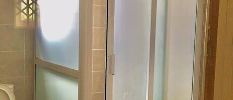 Un baño con cabina de ducha de vidrio, inodoro y ventana con celosía decorativa.