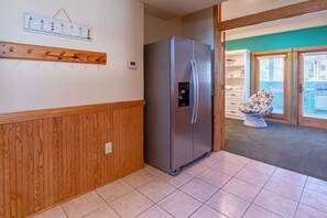 Cozinha com geladeira de aço inoxidável, painéis de madeira, porta-roupas e vista para um quarto com cadeira e estantes.