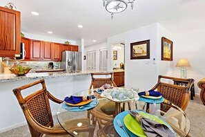 A dining area with a glass table, wicker chairs, and a kitchen in the background. - Spacious 1bd, ground floor, lanai, walk to beach (Kihei)