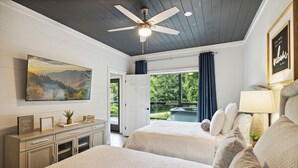 A bedroom with two beds, a ceiling fan, a painting, a desk, and a view of the outdoors. - New Modern Cabin:  Fire Pit, Hot Tub, WiFi, High-End Decor and Amazing Game Room (Gatlinburg)