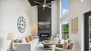 A modern living room with a fireplace, a ceiling fan, and a large window offering a view of the outdoors. - New Modern Cabin:  Fire Pit, Hot Tub, WiFi, High-End Decor and Amazing Game Room (Gatlinburg)