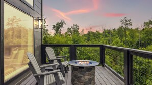 A deck with Adirondack chairs, a stone fire pit, and a view of a forested area. - New Modern Cabin:  Fire Pit, Hot Tub, WiFi, High-End Decor and Amazing Game Room (Gatlinburg)