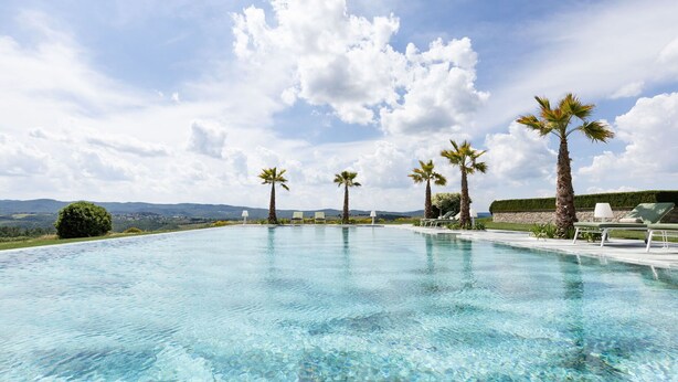 Piscina azul cristalina con palmeras y sillones de descanso, rodeada de jardines y vistas a colinas distantes.