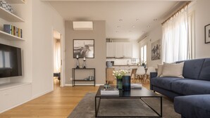 A modern living room with a flat-screen TV, a sofa, and a coffee table. There is a kitchen in the background with white cabinets and a dining area. - Santa Croce Design Apartment R&R (Venecia)