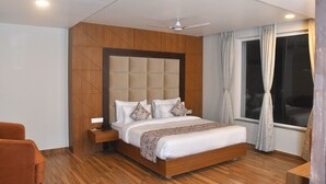 Una habitación de hotel moderna con un cabecero de madera, una cama grande, una mesita de noche, una silla y un ventanal con cortinas.