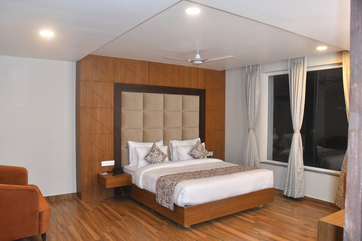 A modern hotel room with a wooden headboard, a large bed, a nightstand, a chair, and a window with curtains.