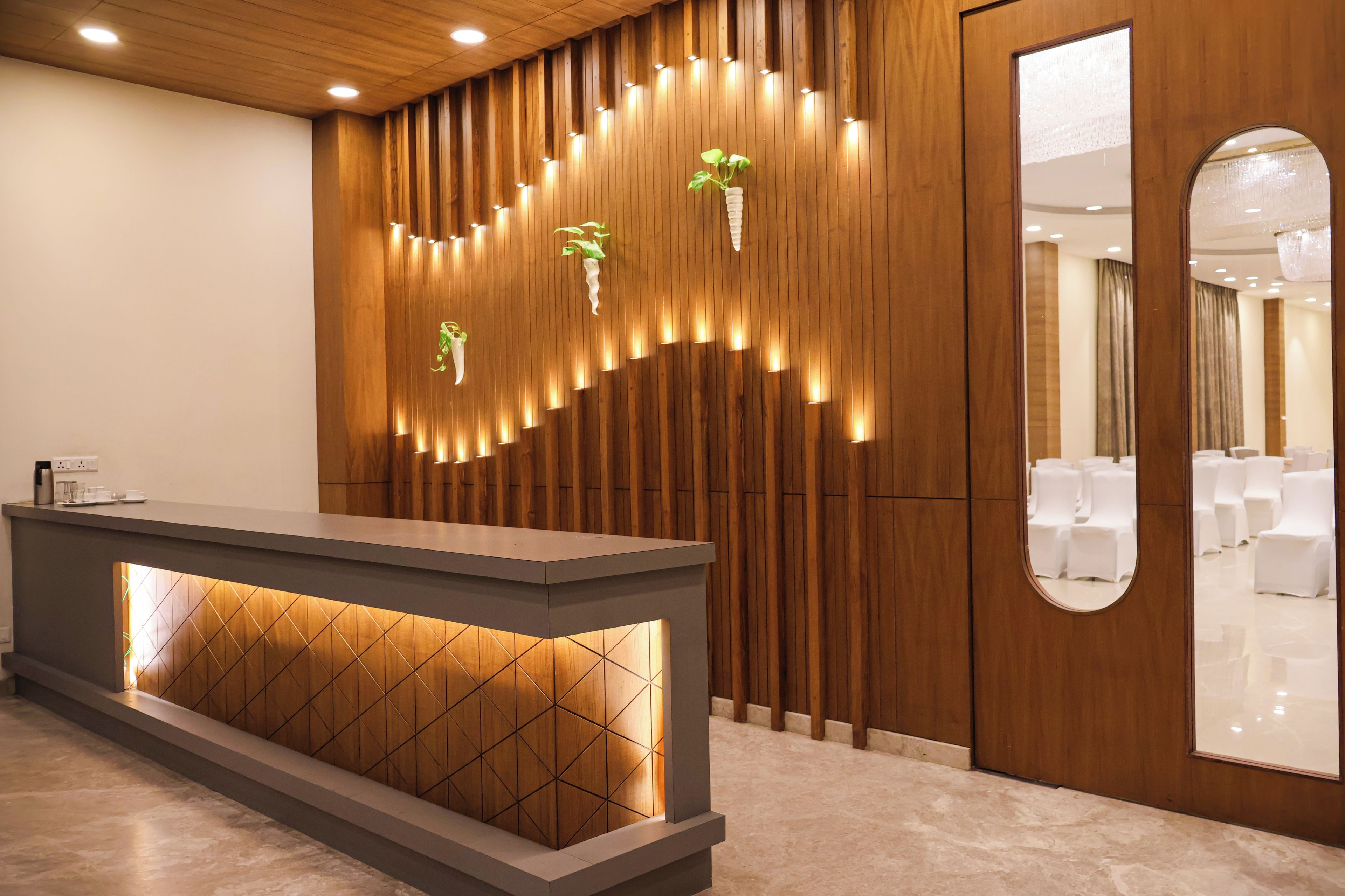 A reception desk with a modern design, a wooden panel wall with integrated lighting, and a mirror reflecting a seating area.