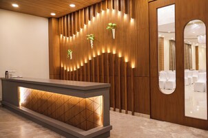 A reception desk with a modern design, a wooden panel wall with integrated lighting, and a mirror reflecting a seating area.