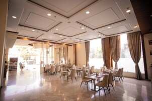 A spacious hotel dining area with multiple tables and chairs, large windows with curtains, and a modern ceiling design.