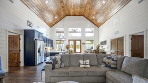 Ein geräumiges Wohnzimmer mit einem großen, grauen Ecksofa, einer Holzbalkendecke und einer Küchenzeile im Hintergrund.