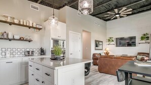 A modern kitchen with white cabinetry, a central island, and pendant lighting. There is a dining area with a black table and a brown leather couch.