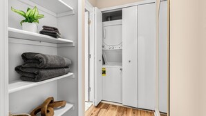 A laundry room with a white washing machine, a mirror, and shelves with folded towels and a plant.