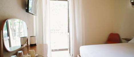 A hotel room with a bed, a bedside table with a mirror, a lamp, and a view of the outdoors.
