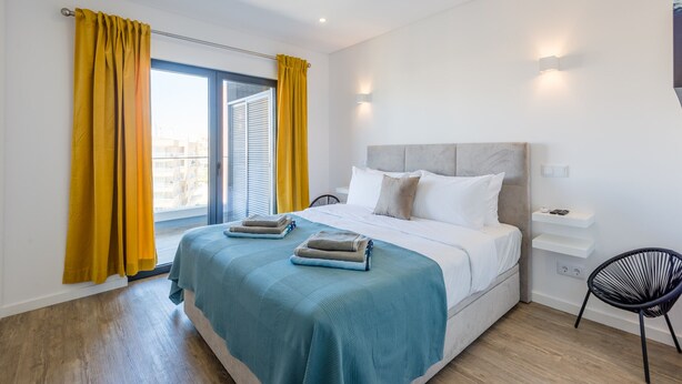 Une chambre moderne avec un grand lit, une table de chevet, une chaise et une vue sur la ville par une fenêtre ornée de rideaux jaunes.
