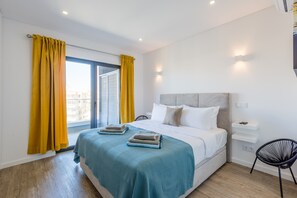 A modern bedroom with a large bed, a nightstand, a chair, and a view of the city through a window with yellow curtains.