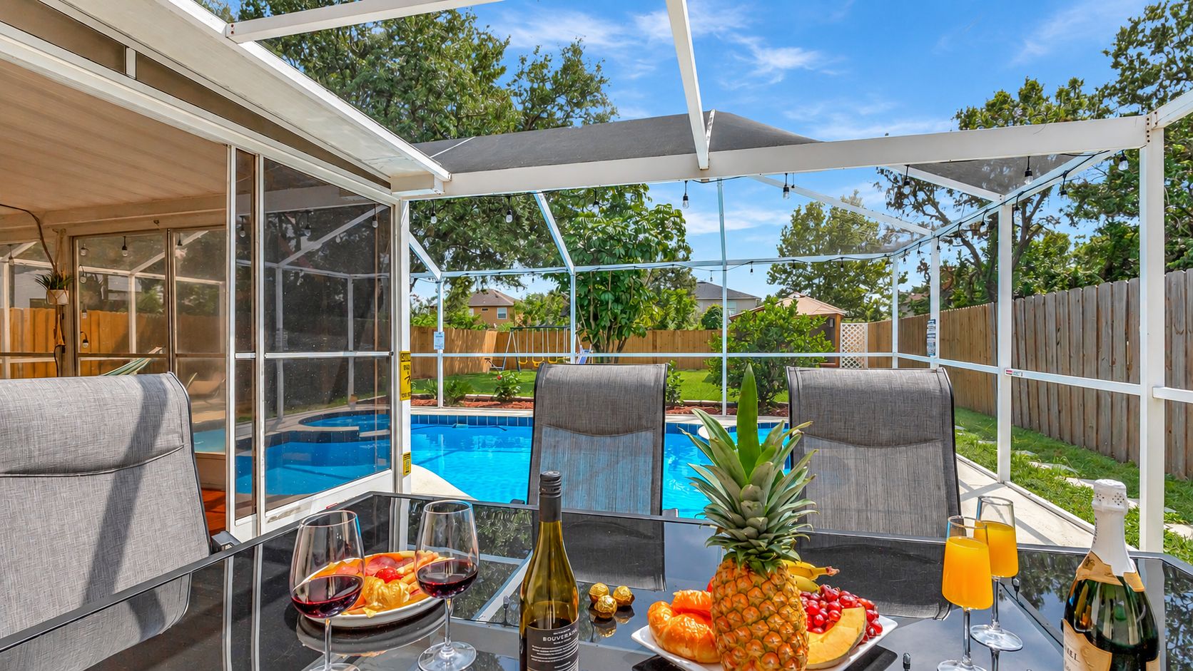 Un patio exterior con cerramiento de vidrio, una piscina y una zona de comedor con comida y bebidas.