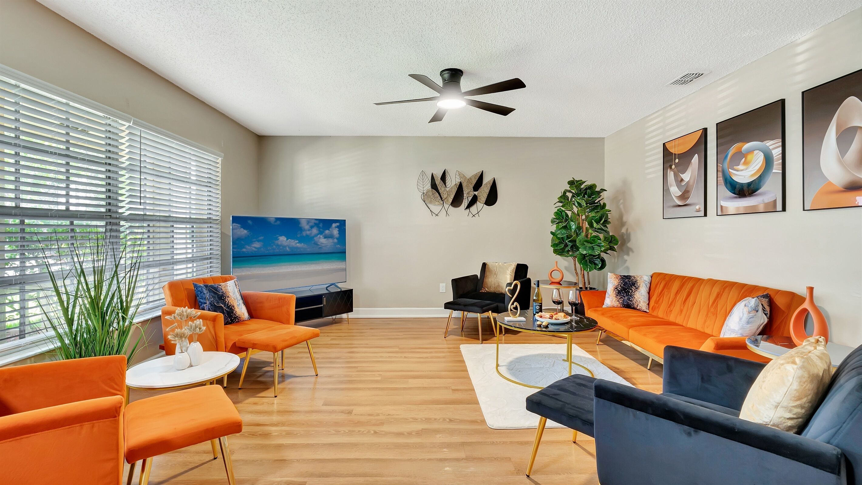 Un salon moderne doté d’un grand téléviseur à écran plat, de canapés orange et noirs, d’un ventilateur de plafond et d’une grande fenêtre avec des stores.