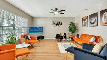 Un salon moderne doté d’un grand téléviseur à écran plat, de canapés orange et noirs, d’un ventilateur de plafond et d’une grande fenêtre avec des stores.