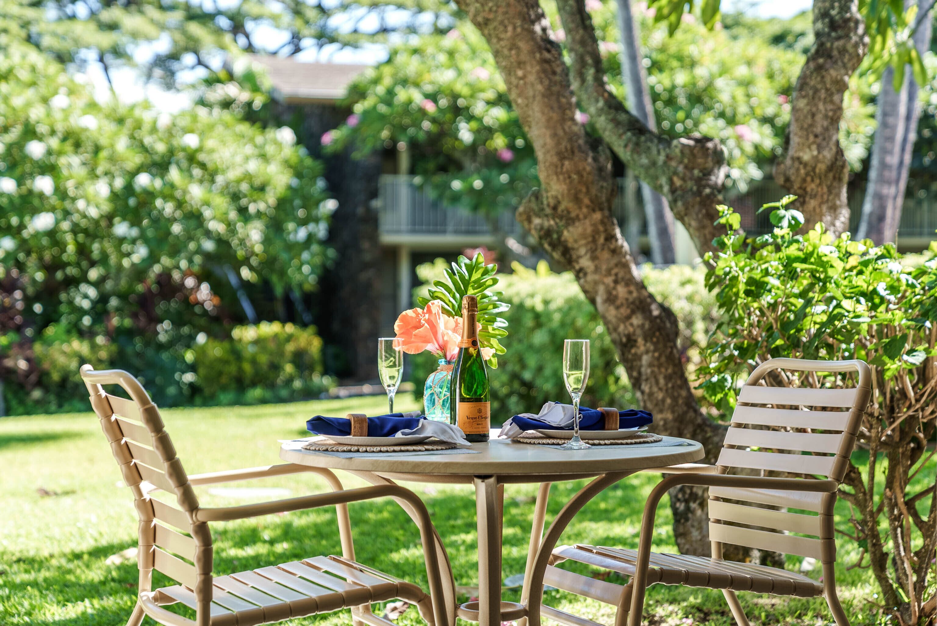 Un espace de restauration en plein air avec deux chaises en osier et une table ronde, agrémenté d’une bouteille de vin, de verres et d’un vase rempli de fleurs.