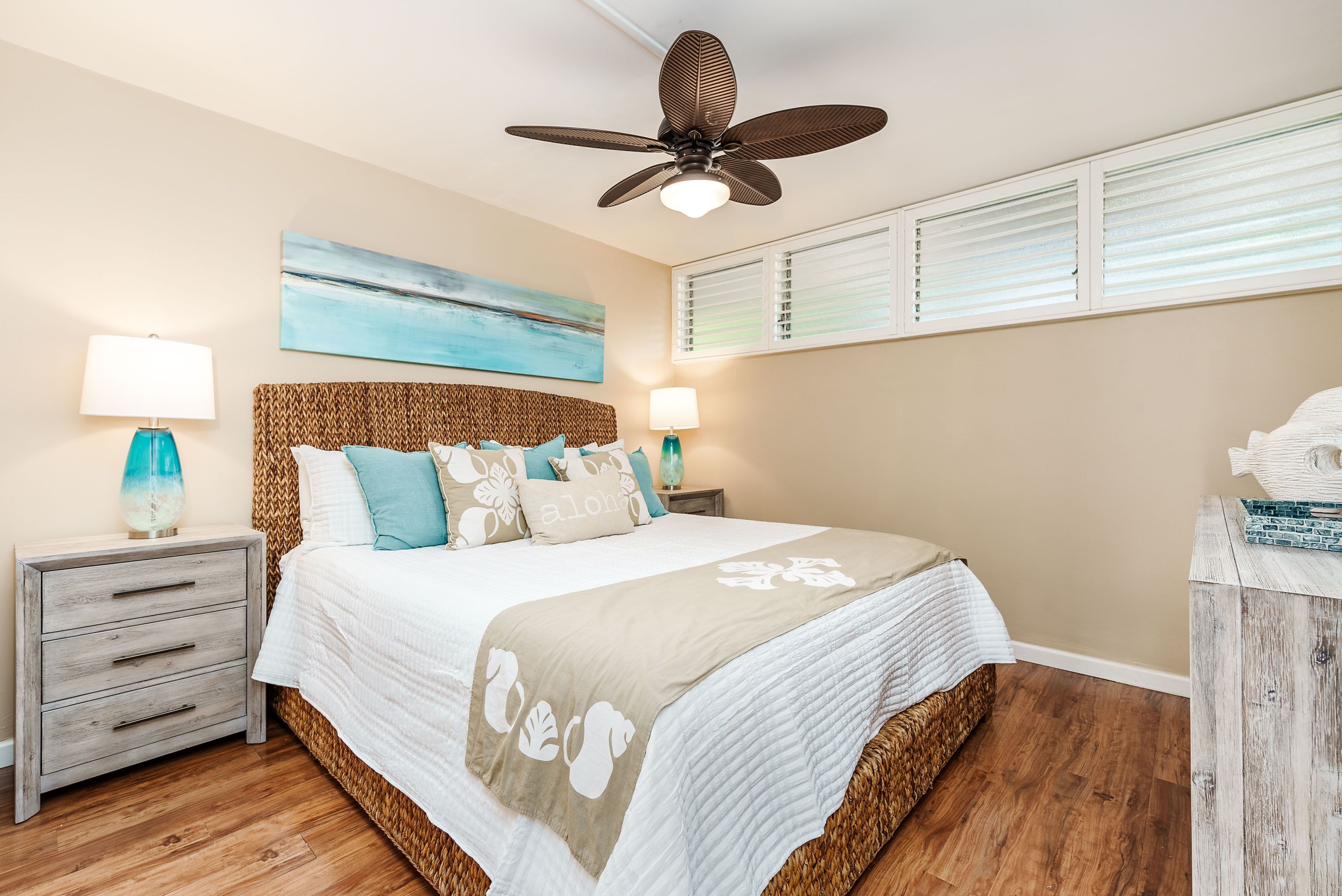 Une chambre à coucher comprenant un lit, deux tables de chevet, un ventilateur de plafond, une fenêtre avec des stores et un tableau accroché au mur.
