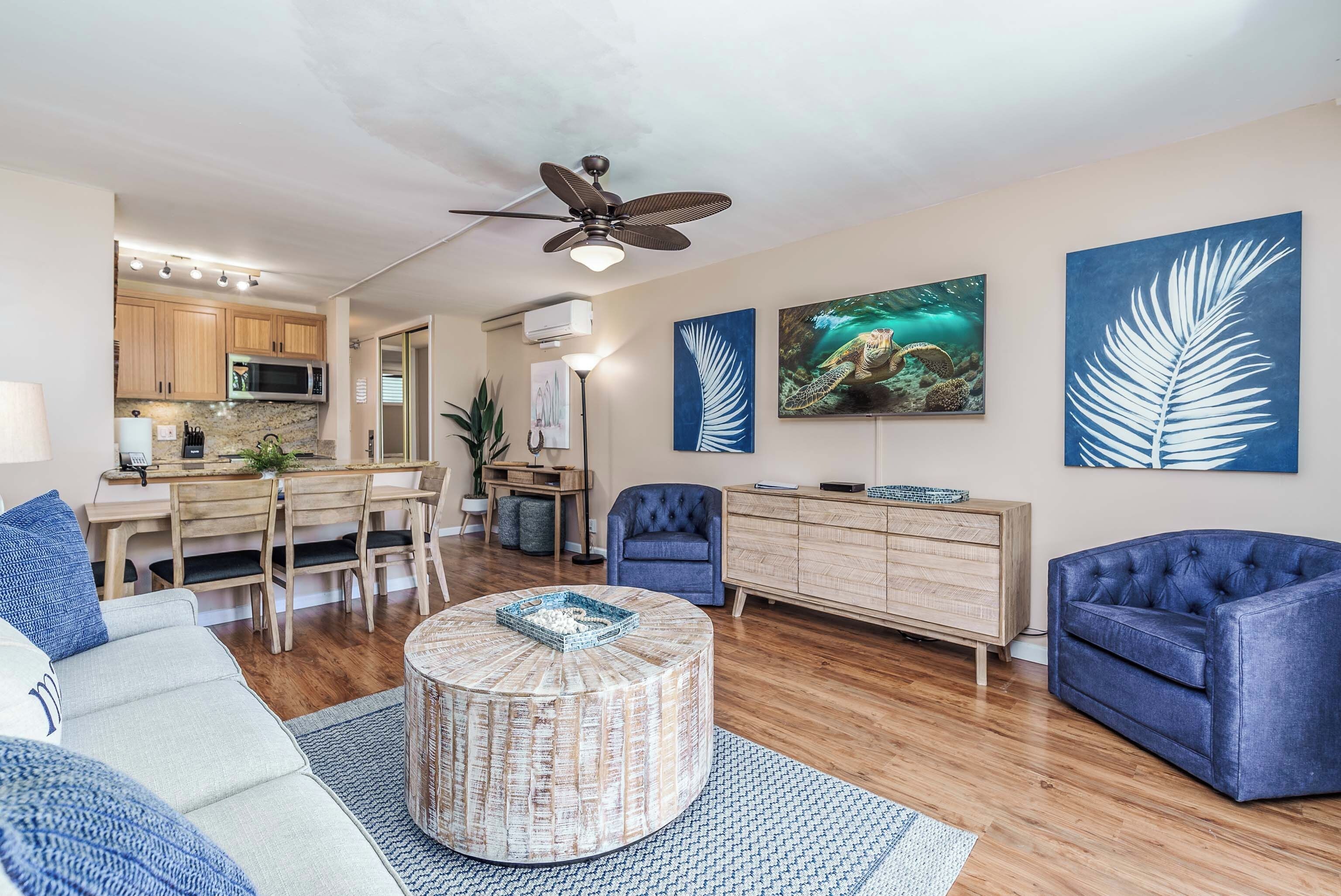 A modern living room with a round coffee table, blue sofas, a dining area with wooden chairs, and artwork on the walls.